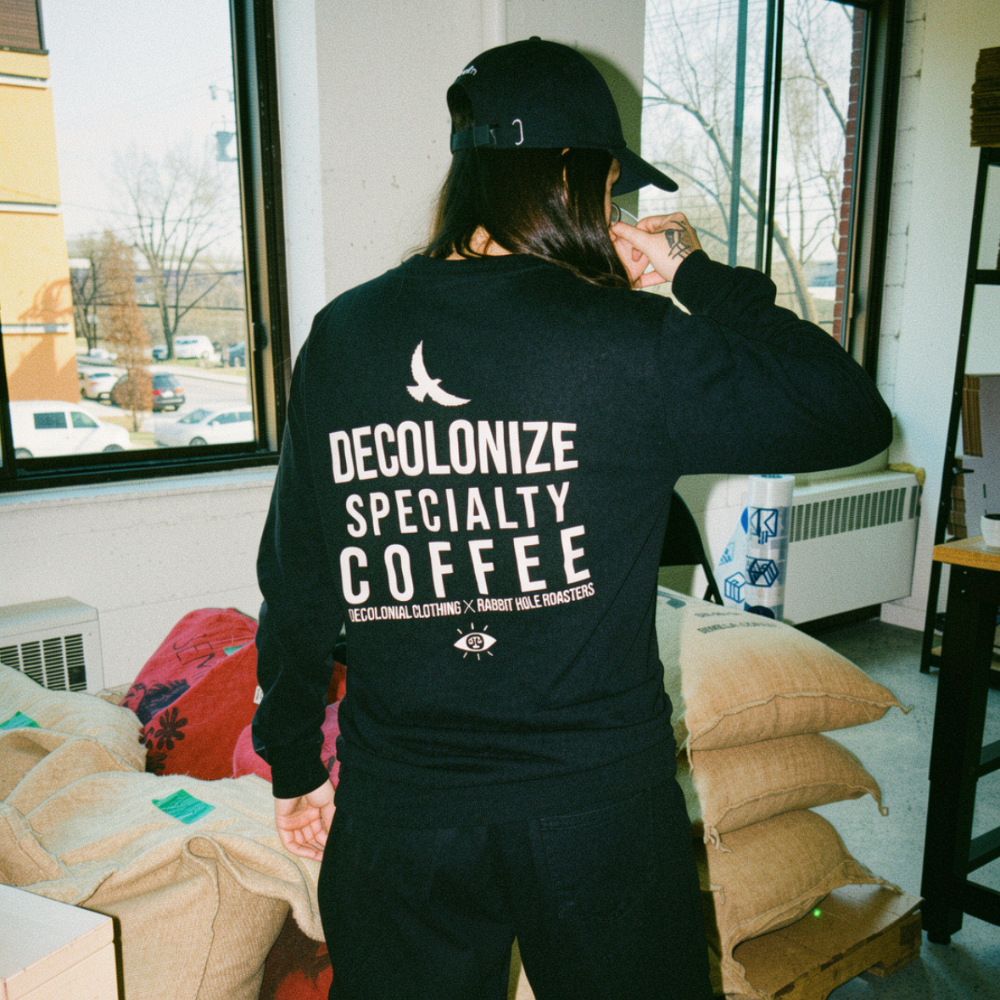 Veronica Ortiz Rodriguez is wearing a black long sleeve tshirt with 'Decolonize Specialty Coffee' printed on the back, by Decolonial Clothing. She casually stands by a window, facing jute coffee bags and sipping on coffee.